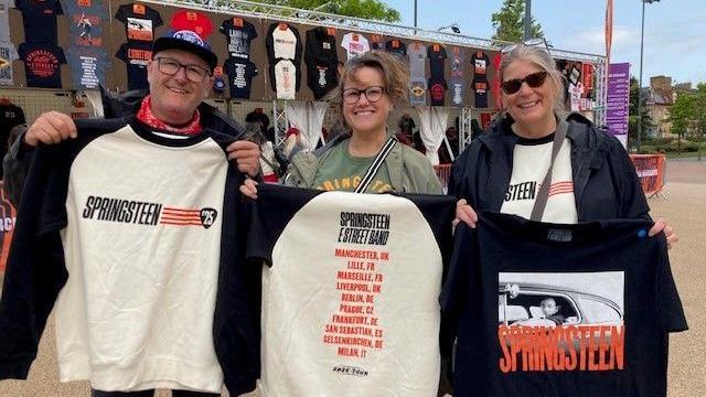 Gareth has glasses and is wearing a baseball cap. Jo is wearing glasses and a green top. Sheryl is wearing sunglasses and a Bruce Springsteen t-shirt. All are holding up Springsteen T-shirts bought from the merchandise stall behind them.