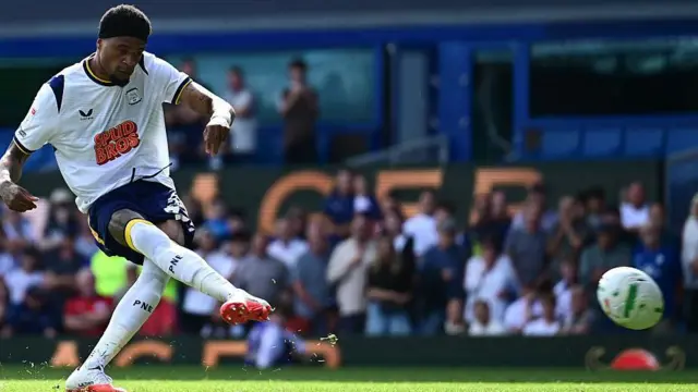 Preston North End footballer Delano Burgzorg wearing a predominantly white kit with blue shorts and yellow trim and hitting the winning penalty with his right foot using a white and green ball in a shootout during the 2026/27 Carabao Cup first round match between the Championship club and League One team Huddersfield Town at Goodison Park in Liverpool