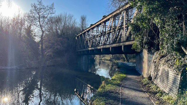 A steel truss bridge over a canal in Wolverhampton on a bright day, with a blue sky and a large flare of light in the top-left corner of the photo. Trees line the left-hand side of the canal and are reflected on the water. There is a footpath on the right side.