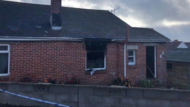 A property with a missing window. The bricks around the outside of the window are blackened and the drainpipe above it has melted