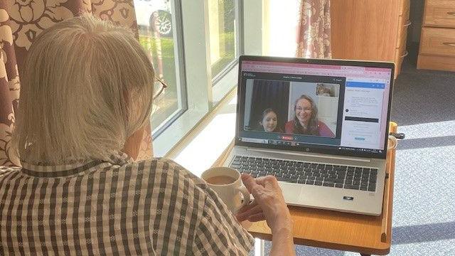 An elderly lady can be seen reaching for a cup of tea whilst the screen on a laptop in front of her shows a lady and a girl who are engaged in a video call with the elderly lady.