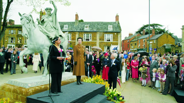 Surinder Dhesi, mayor of Banbury in 2005, gives a speech besides Princess Anne in front of a crowd of people at the unveiling of the Fine Lady on a White Horse statue. A lot of people have gathered with flowers. It is a cloudy day.