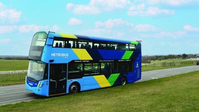 A metrobus hydrogen bus can be seen. It is coloured blue, yellow and green. It is on a quiet road. Either side of there are green fields.