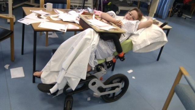 Jamilla Blake as a child in a hospital ward in her reclining wheelchair. She is wearing a hospital gown and smiling at the camera.
