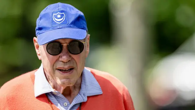 A close up of Michael Eisner wearing a blue cap with the Portsmouth club badge in the centre, tinted, round sunglasses and a blue and white striped shirt underneath an orange jumper