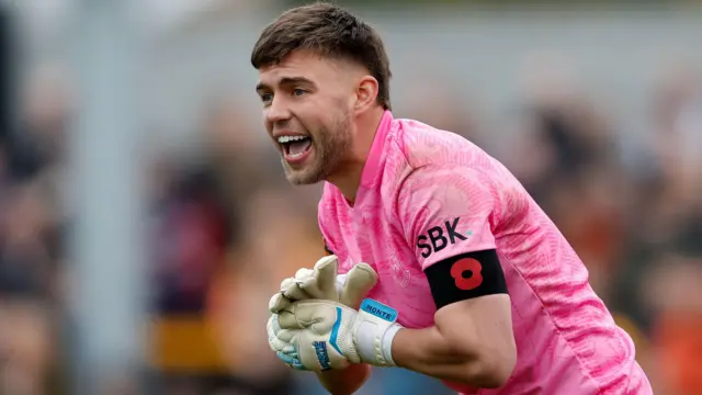 Goalkeeper Jack Sims, pictured in the pink goalkeeping strip at Sutton United, shouts and claps hands.