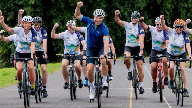 An image of Sir Chris, on his bike, cycling around a park with a large group of cyclists in Tour De 4 cycling tops. They are all punching the air and smiling. He is in the centre, all the cyclists are wearing helmets.