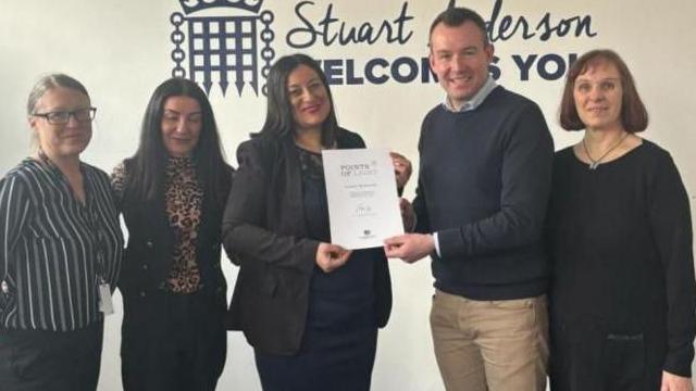 A man smiles as he presents a woman with a certificate. Two other women are standing to her left and another woman is to the man's right.