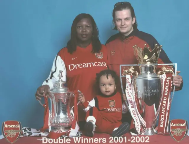 A picture of a man and a woman and a small child sat in between the Premier League trophy and the FA Cup