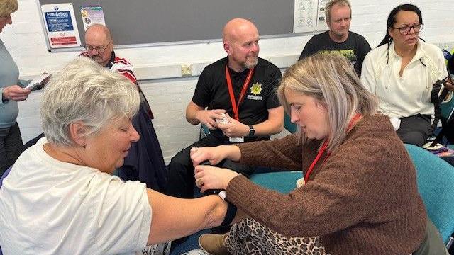 A woman with short white hair wearing a white t-shirt has her arm out. Another woman, with longer silver-blonde hair, a brown jumper an cheetah-print leggings is wrapping a black tourniquet strap around her arm