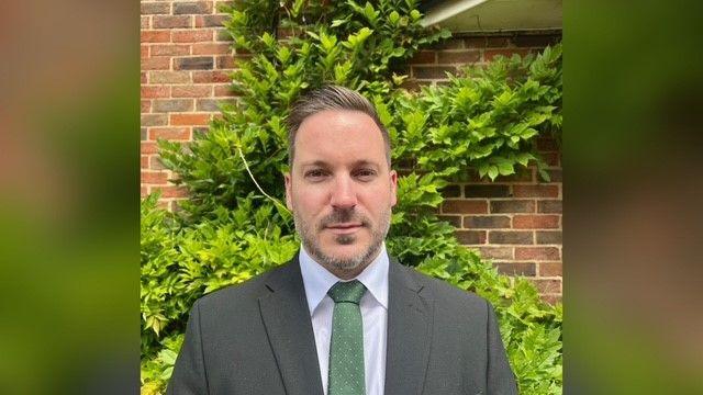 Det Chief Insp James Ansell is standing outside a Surrey Police building. He's got dark blond hair and stubble and is wearing a grey suit and a green tie. There is a brick wall with foliage on it behind him