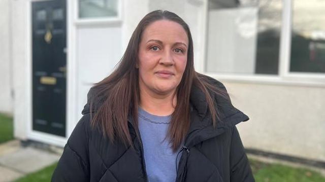 A woman wearing a blue jumper and overcoat stands in front of a house.