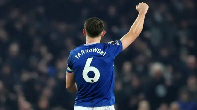The back of James Tarkowski, wearing a blue Everton number six shirt and raising his right fist towards the crowd, which is out of focus in the background