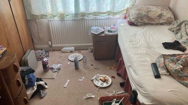 An untidy bedroom, littered with dirty crockery, unmade bedding and sweet wrappers