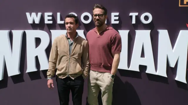 Rob McElhenney and Ryan Reynolds pose in front of a 'Welcome to Wrexham' sign