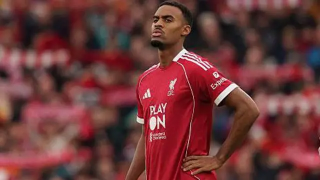 Ryan Gravenberch, wearing Liverpool's red home kit and standing with his hand on his hip during a game, with the Anfield crowd blurred in the background