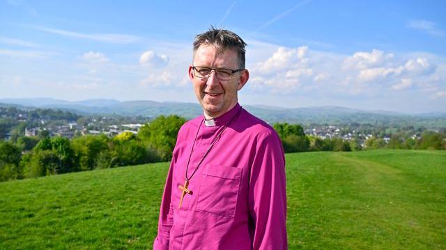 The Right Reverend Rob Saner-Haigh in a bright pink cleric's top and wearing a large crucifix. He is wearing glasses and smiling while standing on a green grassy hill above a town. There are blue skies.