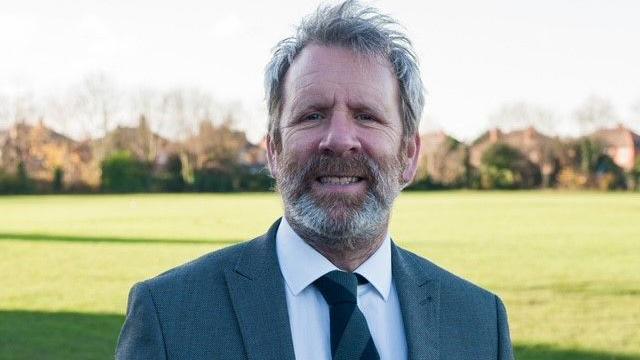 A middle aged man in a grey suit and tie smiles at the camera. He has grey hair and a grey beard and is standing on a grass field which is bathed in sunshine.