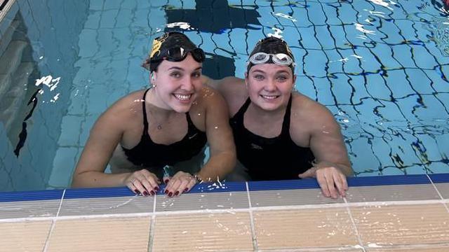 Two women in black swim caps and goggles and black swim costumes stand in a pool and look at the camera smiling.