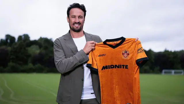 A smiling Cesar Peixoto wearing a grey suit jacket and white t-shirt holds an old gold Wolves home shirt while standing at the club's training ground