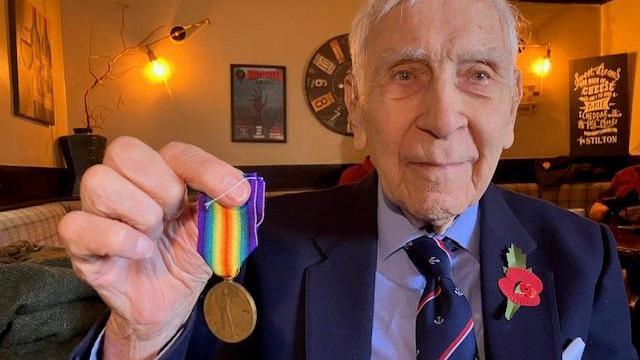 A man in a dark blue suit and light blue shirt with tie holding up a medal by its multi-coloured ribbon