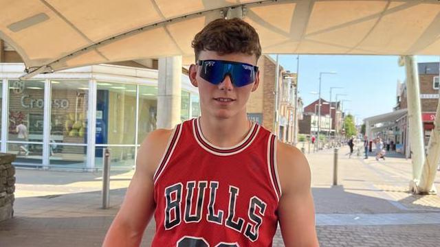 A young man with curly brown hair and sunglasses wearing a red vest with the word "BULLS" on it. He is stood in front of a high street on a sunny day.