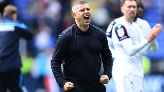 Steven Schumacher, wearing a long sleeve blue top, clenches his fists by his sides and shouts to celebrate Bolton's win over Preston 