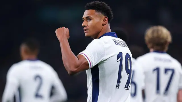 Ollie Watkins, wearing England's white home shirt, clenches his fist towards supporters after scoring a goal. Anthony Gordon and Ezri Konsa are blurred in the background.