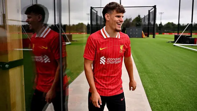 Milos Kerkez smiling at Axa Training Centre in a Liverpool shirt