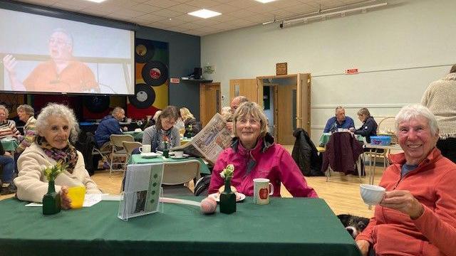 A community brunch with two women and a man smiling at the camera, behind them is other tables of people in a community hall with a powerpoint video of a man speaking.