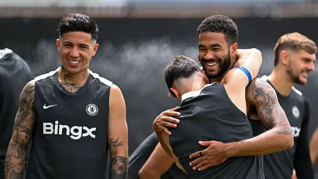 Enzo Fernandez and Reece James wear black Chelsea training vests and grin at the camera as another player with his back to the photographer puts his arms around James' neck