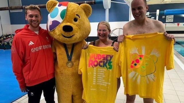 Dan and Sophie wearing swimwear and holding up yellow Pudsey T-shirts, posing for a photo with Pudsey Bear and Kieran, who is wearing a red 'Great Britain' hoodie. They're inside a leisure centre and there's a swimming pool behind them.