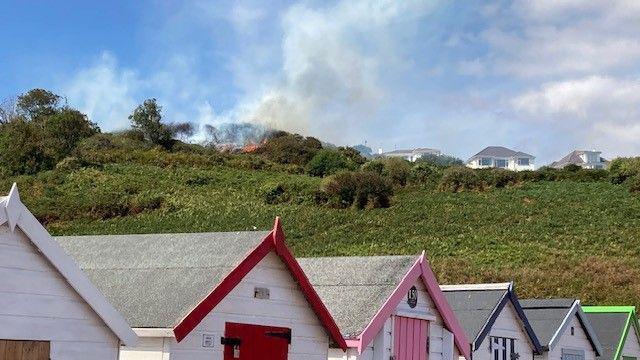 There is a fire at the top of the hill. Along the bottom are beach huts which are different colours. There are also houses at the top of the hill on the right.