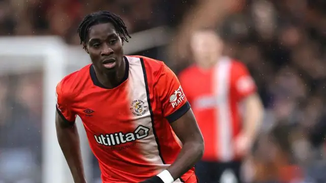 Elijah Adebayo in match action for Luton wearing an orange playing shirt with a white vertical stripe from the left shoulder down the front of the shirt