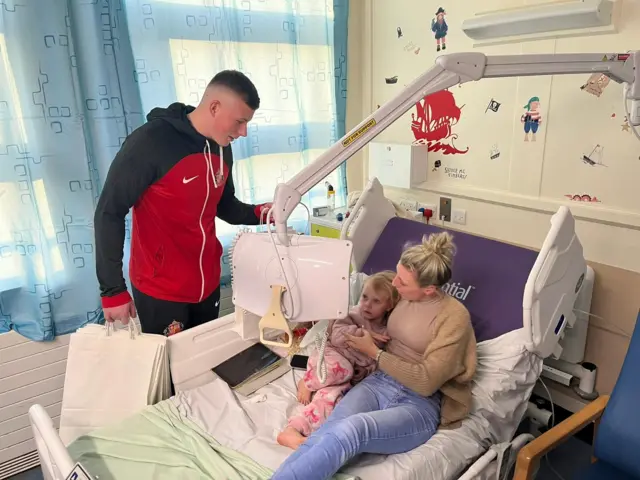 Anthony Patterson handing a bag of presents to a young girl and her mam while they lie in a hospital bed