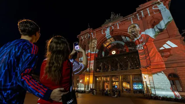Two Arsenal supporters take a photo of the Budapest-Keleti train station with a projection of Martin Odegaard and Gabriel, the Adidas logo and the words 'YOU GOT THIS'