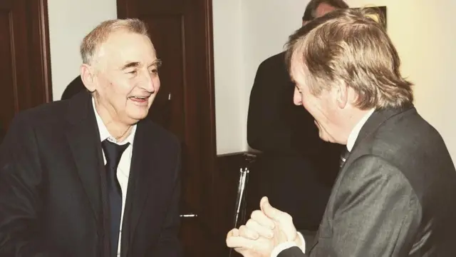 A photo of Tony Parkes (left), wearing a black suit and tie, talking to Sir Kenny Dalglish (right)