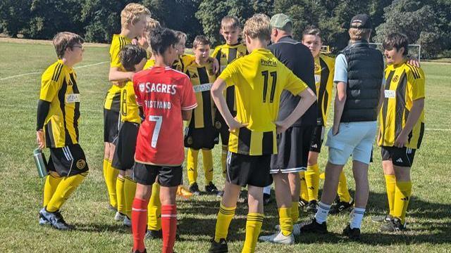 Teenage football players on the football pitch standing in a huddle. Most of them have their backs to the camera, wearing their yellow and black shirts.