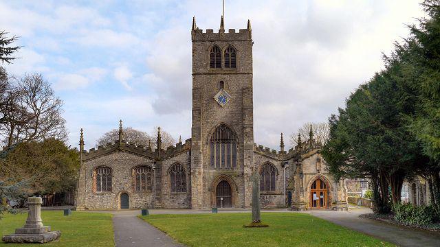 The front face of the Grade I listed Church of Holy Trinity in Kirkland, Kendal. It has several large stained glass windows. It is a stone-coloured and a large impressive building next to trees.