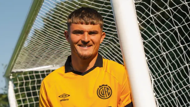 Callum Perry poses against a football goalpost in an amber Cambridge United home jersey.