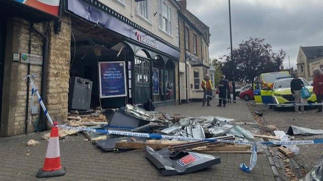 Olney High Street, showing a damaged Nationwide, with material on the floor and police tape, a police car, people standing around and a tree in the distance.