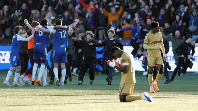 Crystal Palace's Marc Guehi kneels on the pitch as Macclesfield F.C. players celebrate