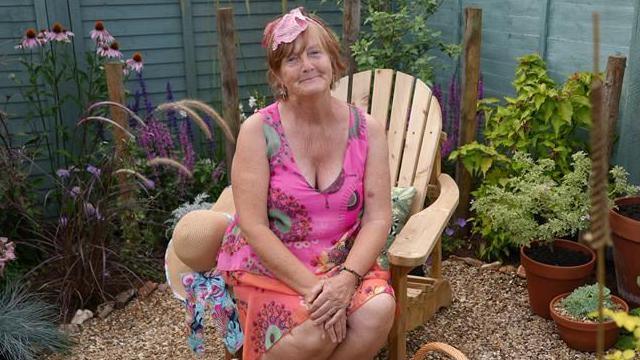 Hillary-Fay Mellor, a woman who is sitting on a wooden chair surrounded by plants and flowers in a garden display. She is looking directly at the camera and smiling. She is wearing a pink and orange patterned dress and has a sun hat on the chair next to her.