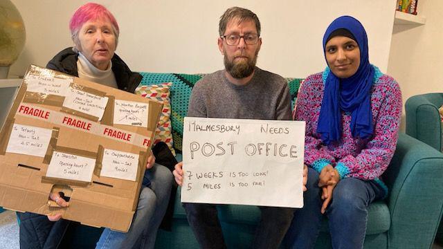 Fran Vandelli, Mubashir Akhtar and Jumana Akhtar. They are all sitting on a turquoise sofa. Fran has short pink hair and is wearing a cream turtle neck with a black coat and blue jeans. She is holding a cardboard sign with six pieces of white card on it, each with a note about the distance to other post offices. Mubashir is sitting in the middle. He has glasses, short dark hair and a beard. He is wearing a grey jumper and is holding a white sign reading "Malmesbury Needs Post Office 7 weeks is too long! 5 miles is too far!" Jumana is sitting next to him. She is wearing a blue hijab and a purple jumper. All of them are looking at the camera. 