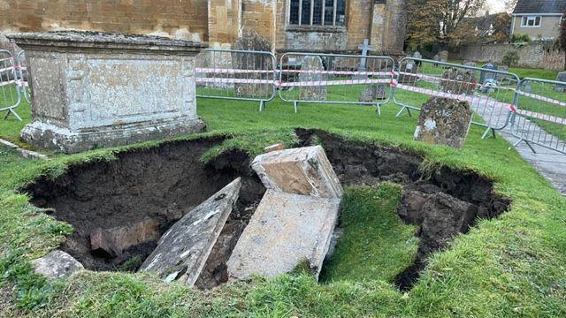 A wide shot from a different angle, showing the large hole which has suddenly collapsed, very near to the surrounding graves. The church is in the background and there are metal barriers securing this section of the graveyard.
