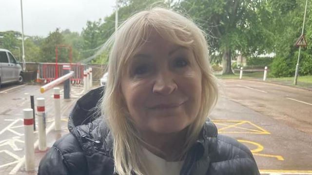 A blonde woman smiling at the cameras. She is wearing a black coat and there is a car park behind her.