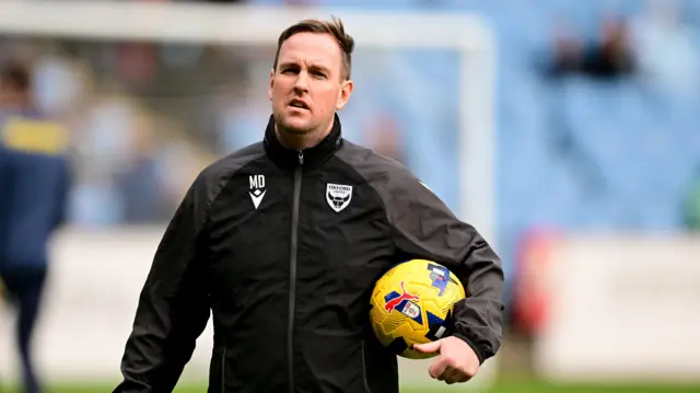 Mike Dodds holding a football under his right arm to his body, wearing an Oxford United black coat while standing on a pitch during a pre-game warm-up