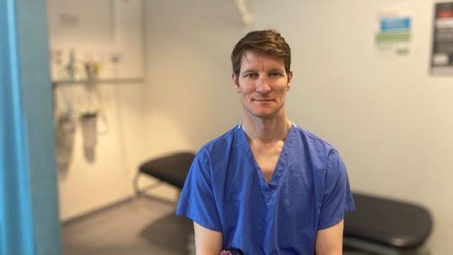 Tim Lawrence is wearing blue medical scrubs and sat in a clinic room