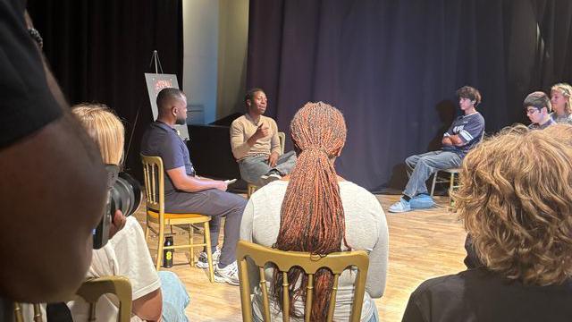 David Jonsson is sat on a chair with a semi circle of young people around him also sat on chairs.There are 8 young people in the image, they are all sat in a room with black curtains covering all the walls and a wooden floor.