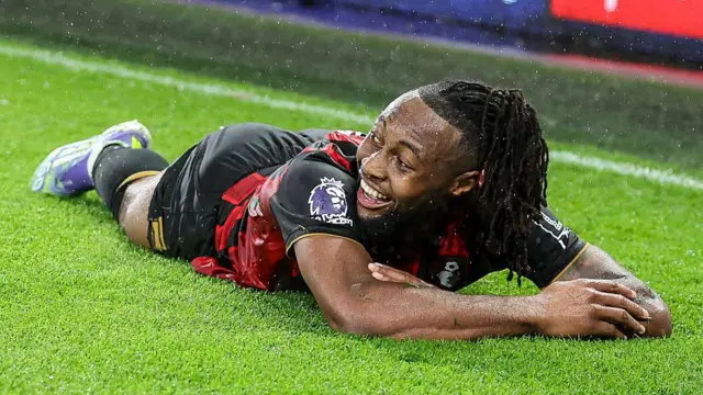 A smiling Antoine Semenyo, wearing Bournemouth's red and black shirt and black shorts, lies on his front on the pitch after scoring a goal
